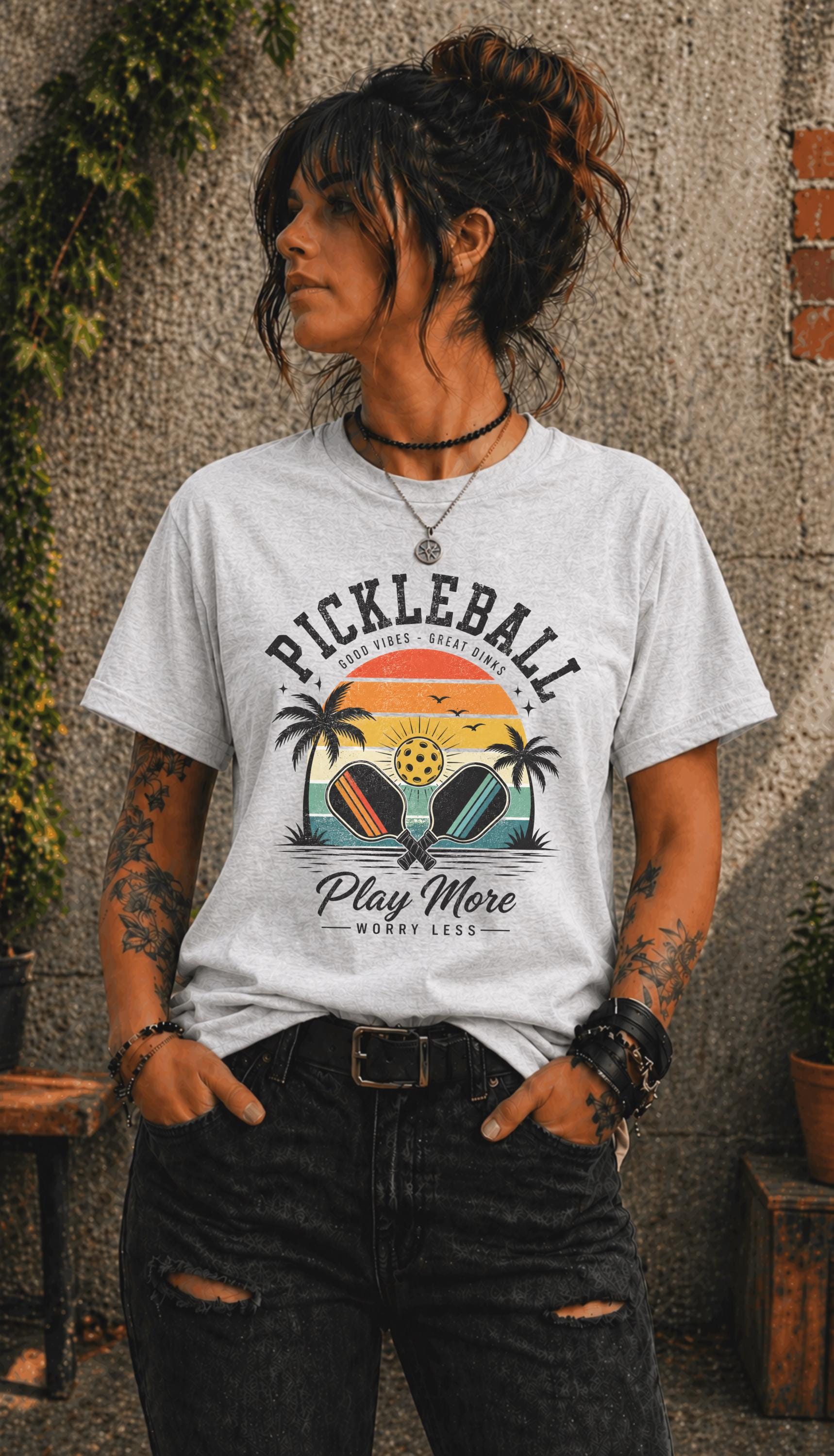 Woman wearing a Pickleball Play More Worry Less graphic t-shirt featuring retro paddles, tropical palm trees, vintage sunset artwork, and coastal-inspired pickleball design.