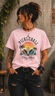 Woman wearing a Pickleball Play More Worry Less graphic t-shirt featuring retro paddles, tropical palm trees, vintage sunset artwork, and coastal-inspired pickleball design.