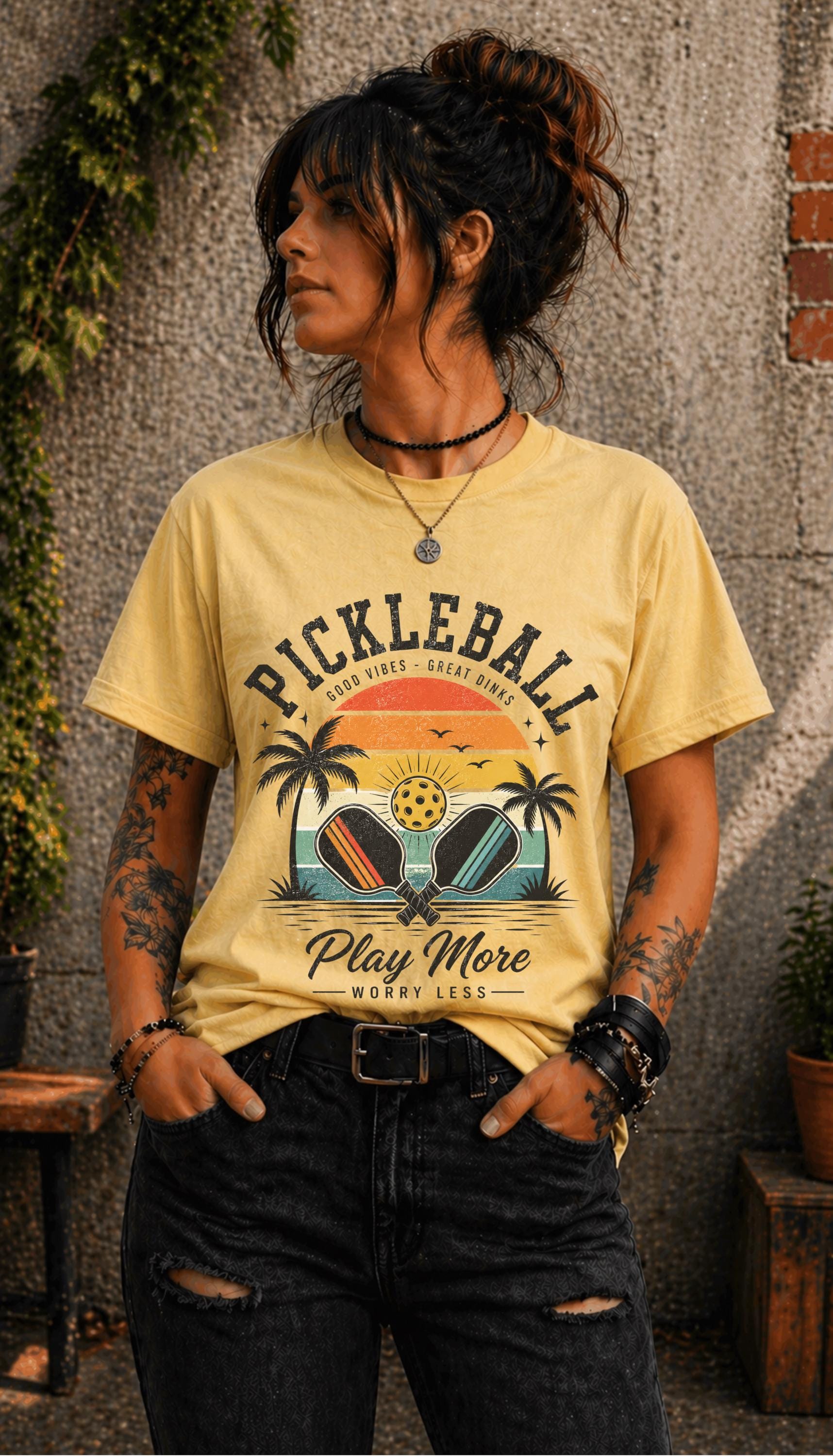 Woman wearing a Pickleball Play More Worry Less graphic t-shirt featuring retro paddles, tropical palm trees, vintage sunset artwork, and coastal-inspired pickleball design.