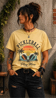 Woman wearing a Pickleball Play More Worry Less graphic t-shirt featuring retro paddles, tropical palm trees, vintage sunset artwork, and coastal-inspired pickleball design.