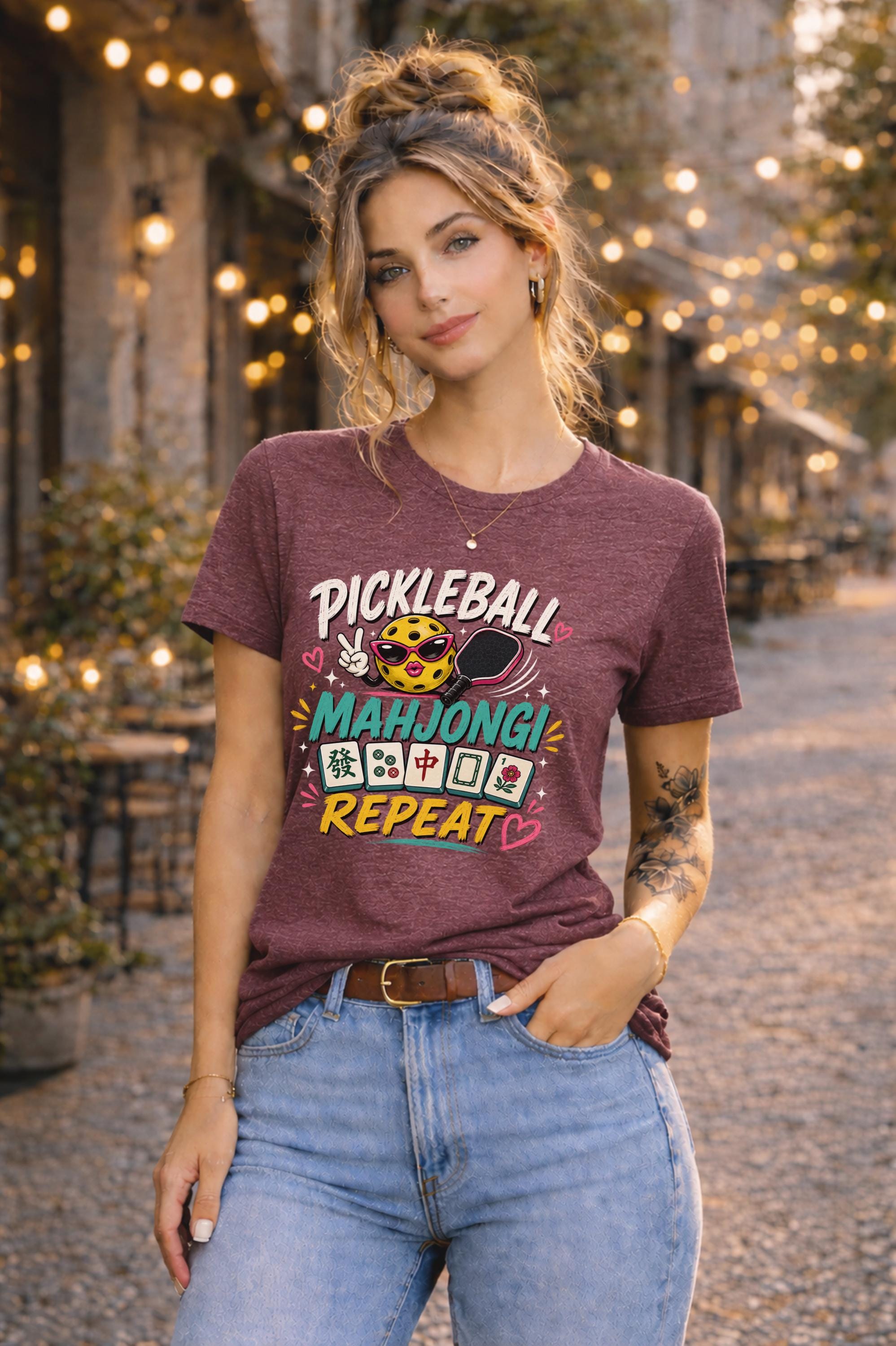 Woman wearing a graphic t-shirt featuring a colorful "Pickleball Mahjong Repeat" design with pickleball character, mahjong tiles, hearts, and retro-inspired artwork.