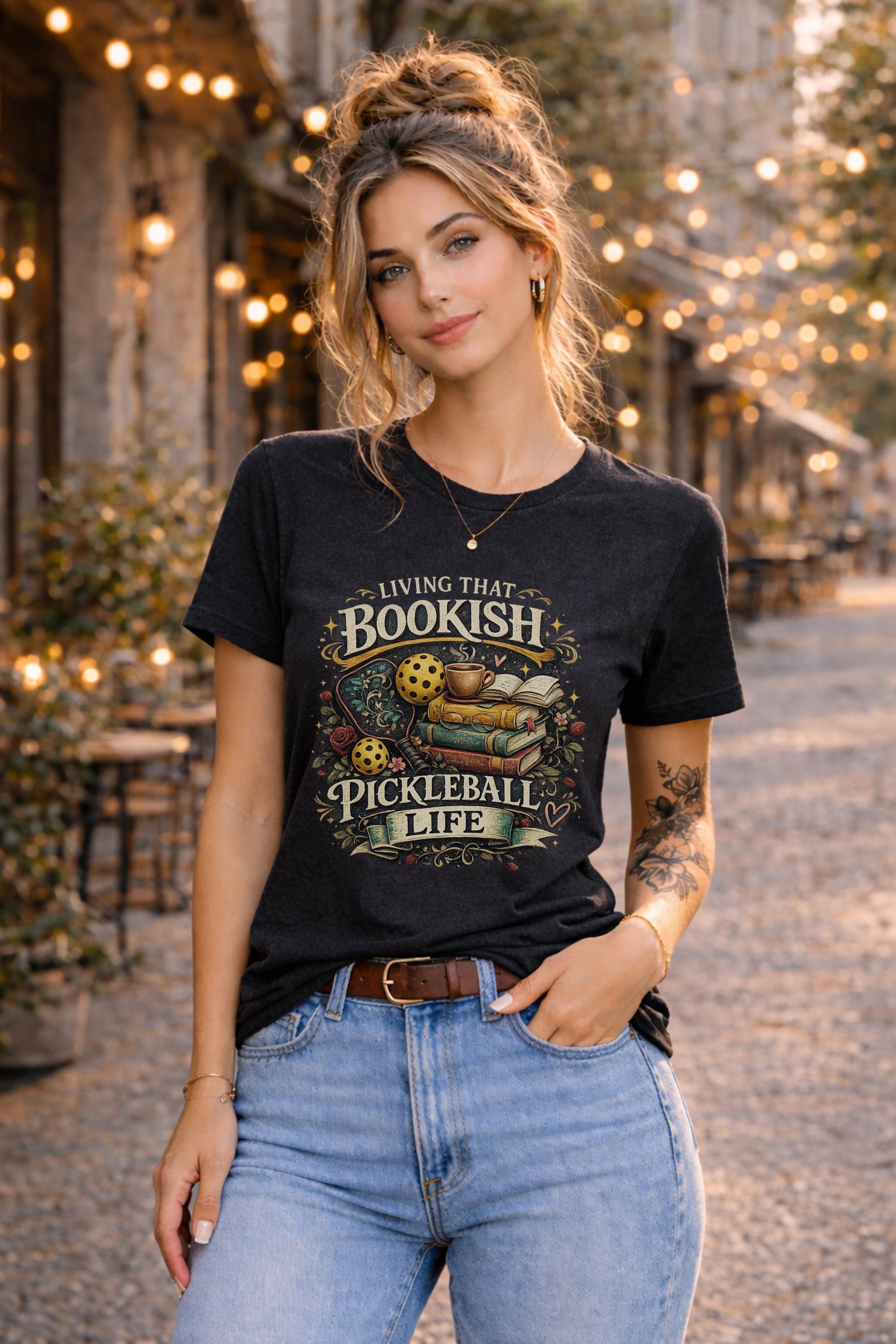Woman wearing a graphic t-shirt featuring a Living That Bookish Pickleball Life design with books, coffee cup, pickleball paddles, floral accents, and vintage-inspired artwork.