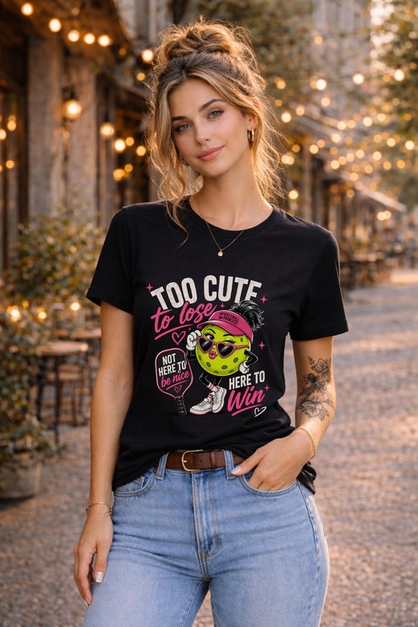 Woman wearing a black Too Cute To Lose pickleball graphic t-shirt featuring a stylish pickleball character with heart sunglasses, pink visor, paddle artwork, and playful court-inspired design.