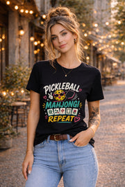Woman wearing a black graphic t-shirt featuring a colorful "Pickleball Mahjong Repeat" design with pickleball character, mahjong tiles, hearts, and retro-inspired artwork.