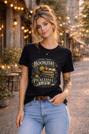 Woman wearing a black graphic t-shirt featuring a Living That Bookish Pickleball Life design with books, coffee cup, pickleball paddles, floral accents, and vintage-inspired artwork.