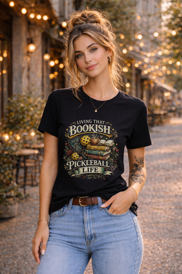 Woman wearing a black graphic t-shirt featuring a Living That Bookish Pickleball Life design with books, coffee cup, pickleball paddles, floral accents, and vintage-inspired artwork.