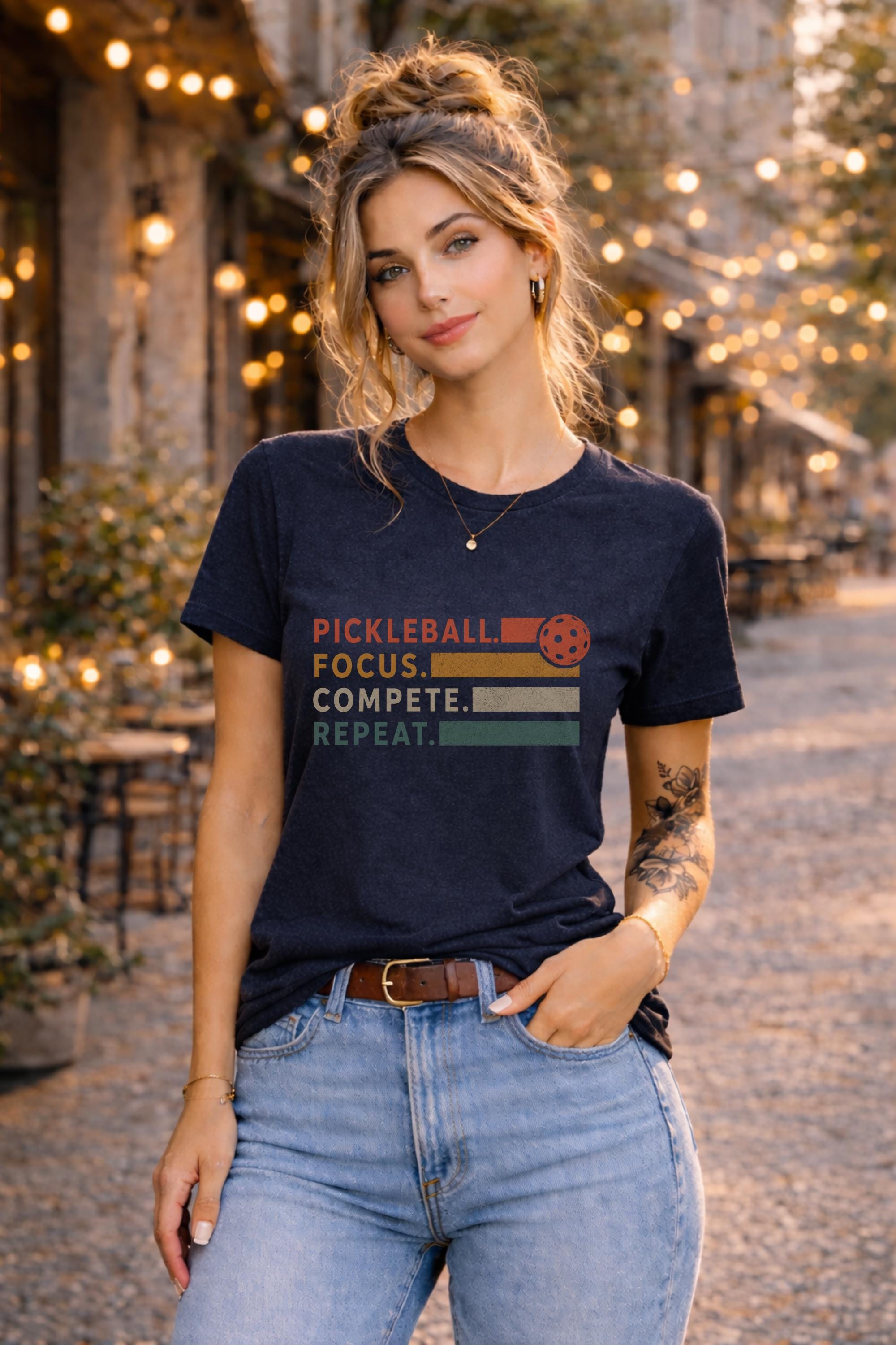 Woman wearing a graphic t-shirt featuring a vintage Pickleball Focus Compete Repeat design with retro stripes, pickleball icon, and motivational athletic typography.