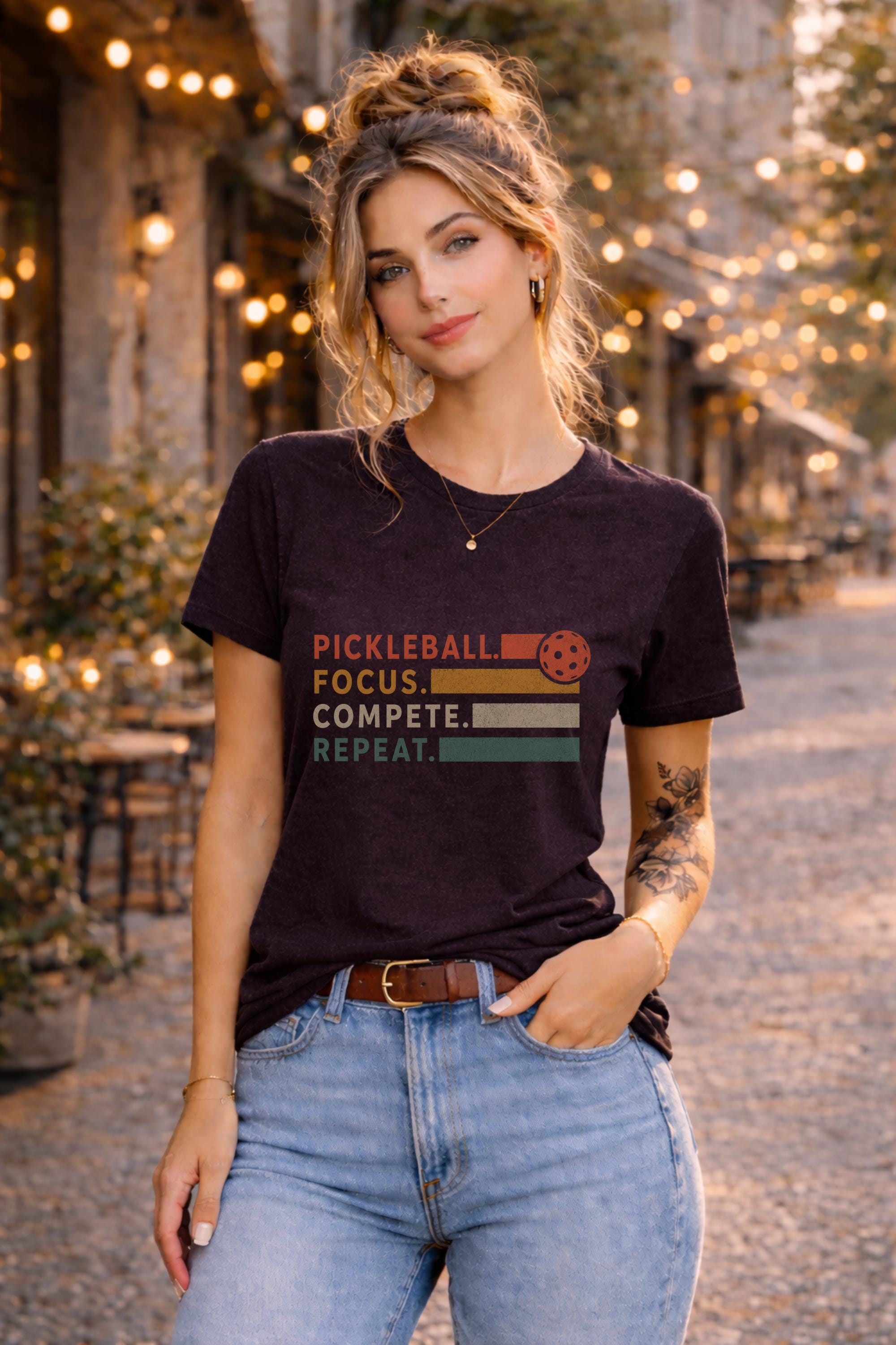 Woman wearing a graphic t-shirt featuring a vintage Pickleball Focus Compete Repeat design with retro stripes, pickleball icon, and motivational athletic typography.