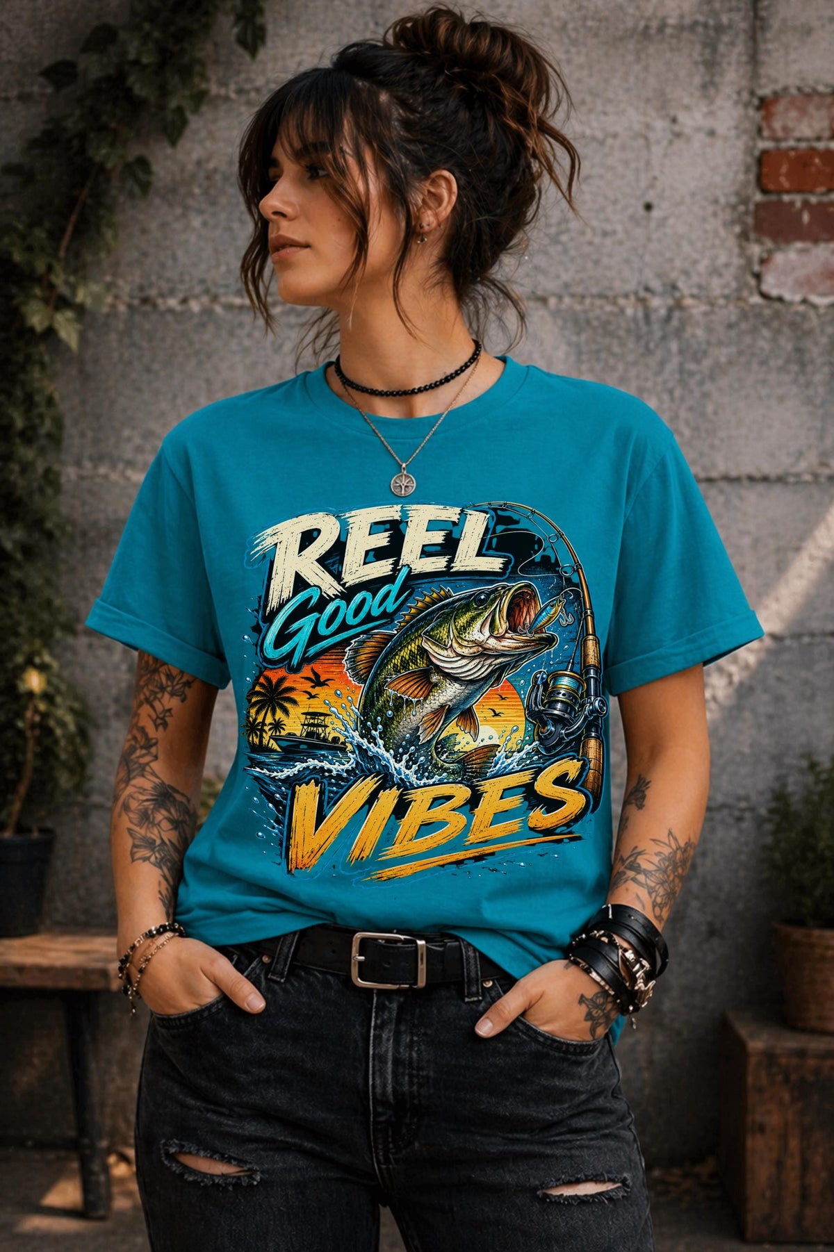 Woman wearing a graphic t-shirt featuring a jumping bass fish, fishing rod, tropical sunset scene, and Reel Good Vibes lettering.