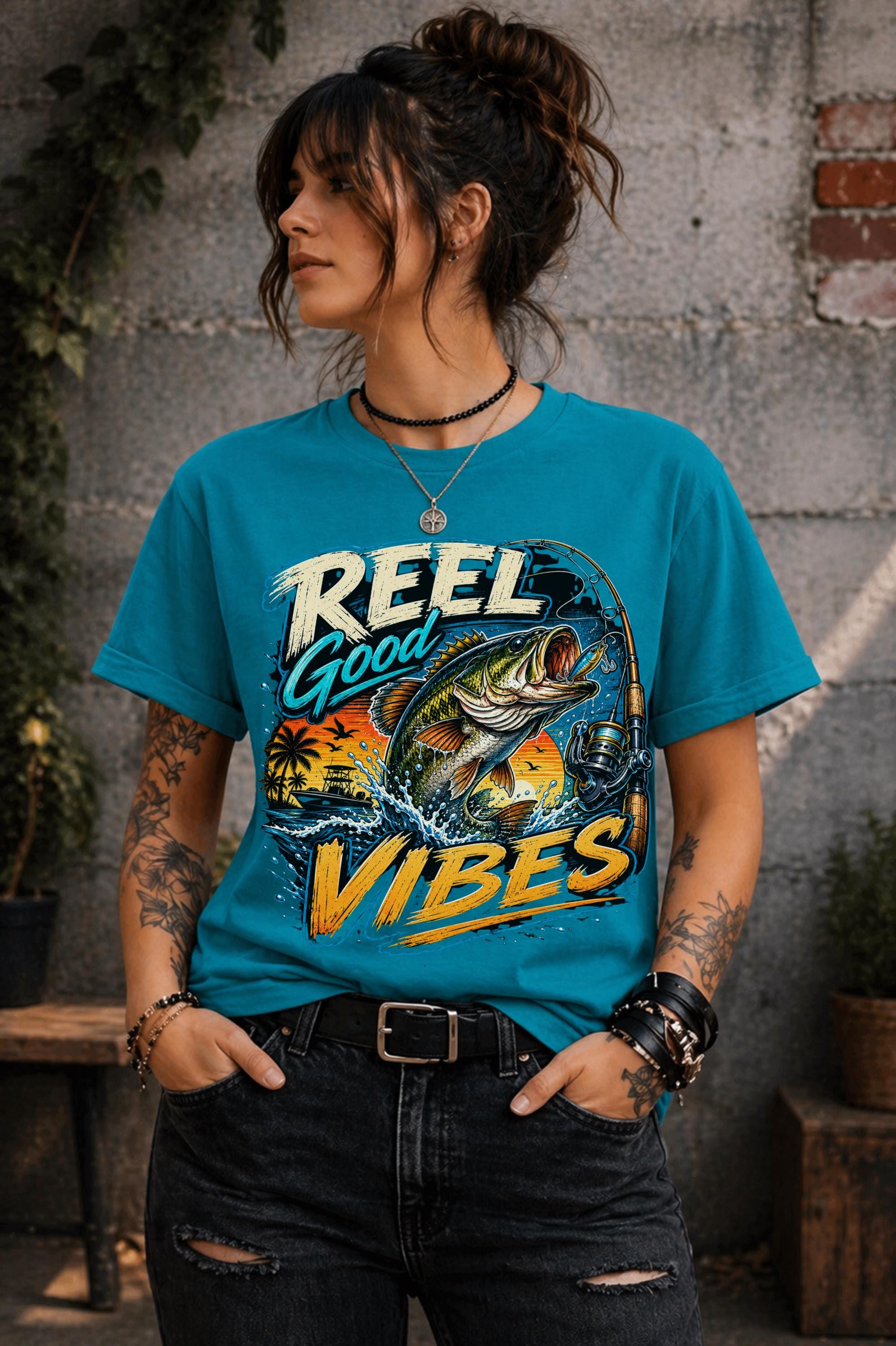 Woman wearing a graphic t-shirt featuring a jumping bass fish, fishing rod, tropical sunset scene, and Reel Good Vibes lettering.