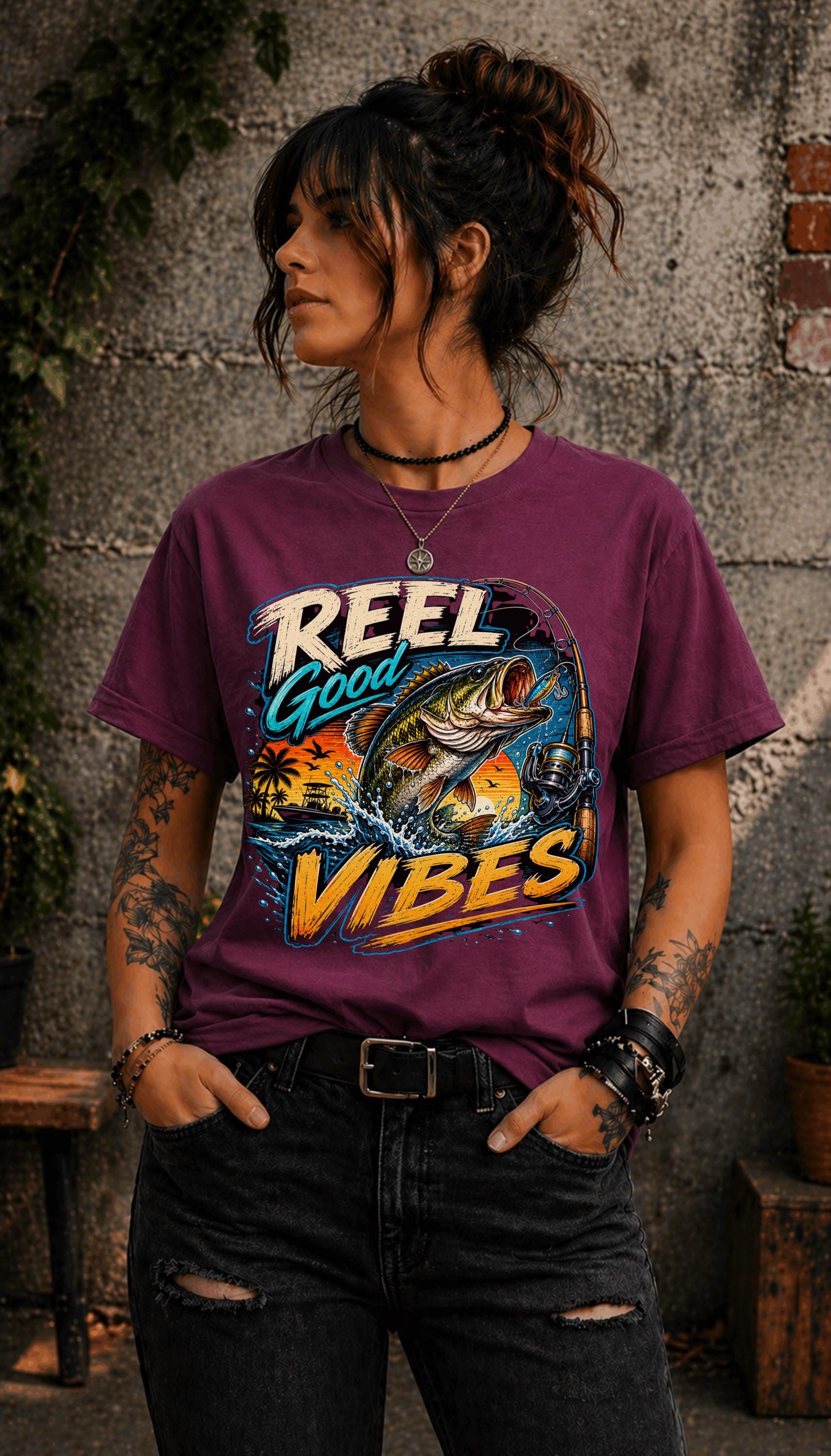 Woman wearing a graphic t-shirt featuring a jumping bass fish, fishing rod, tropical sunset scene, and Reel Good Vibes lettering.