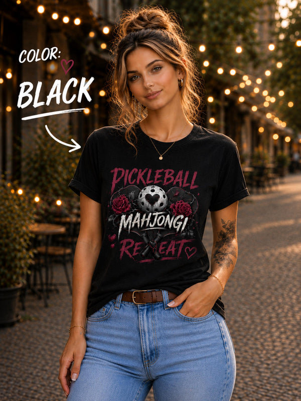 May include: A black short-sleeve t-shirt featuring a graphic print with the text "PICKLEBALL MAHJONG REPEAT". The design includes a central pickleball with a heart cutout, flanked by two red roses, and two crossed pickleball paddles below.