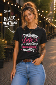 Woman wearing black heather graphic t-shirt featuring the phrase "Probably Late for Something That Doesn't Matter" in pink and white typography with playful heart accents.