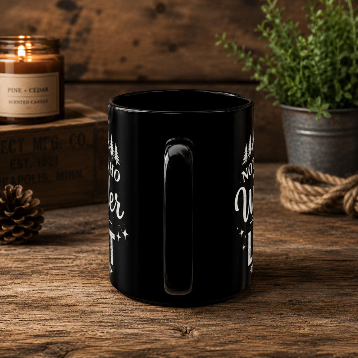 The handle of the Black ceramic mug featuring mountain artwork and the phrase "Not All Who Wander Are Lost" displayed in white typography on a cozy rustic background.