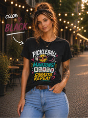 Woman wearing a black graphic t-shirt featuring the colorful phrase "Pickleball Mahjong Canasta Repeat" with a smiling pickleball character, paddle, and Mahjong tiles, styled for casual everyday wear.