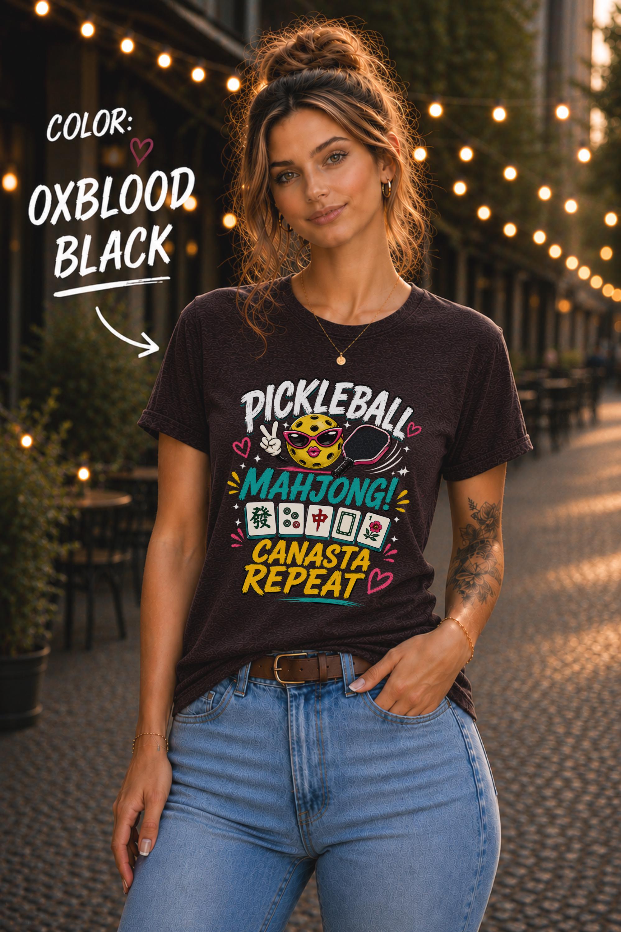 Woman wearing a oxblood black graphic t-shirt featuring the colorful phrase "Pickleball Mahjong Canasta Repeat" with a smiling pickleball character, paddle, and Mahjong tiles, styled for casual everyday wear.