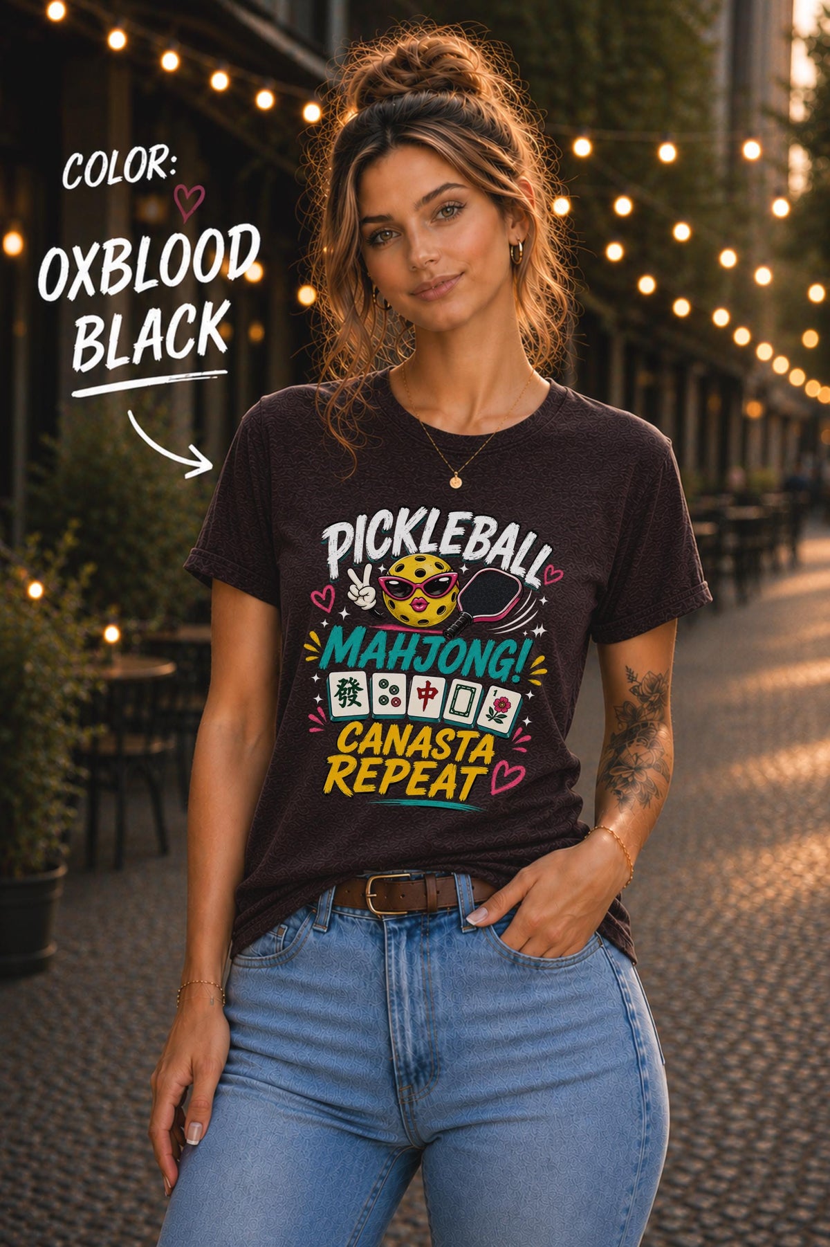 Woman wearing a oxblood black graphic t-shirt featuring the colorful phrase "Pickleball Mahjong Canasta Repeat" with a smiling pickleball character, paddle, and Mahjong tiles, styled for casual everyday wear.