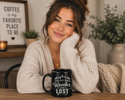 Black ceramic mug featuring mountain artwork and the phrase "Not All Who Wander Are Lost" displayed in white typography on a cozy rustic background.
