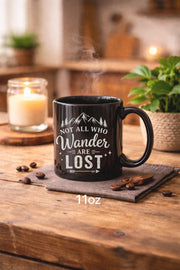 Black ceramic mug featuring mountain artwork and the phrase "Not All Who Wander Are Lost" displayed in white typography on a cozy rustic background.