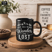 Black ceramic mug featuring mountain artwork and the phrase "Not All Who Wander Are Lost" displayed in white typography on a cozy rustic background.