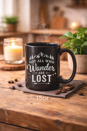 Black ceramic mug featuring mountain artwork and the phrase "Not All Who Wander Are Lost" displayed in white typography on a cozy rustic background.