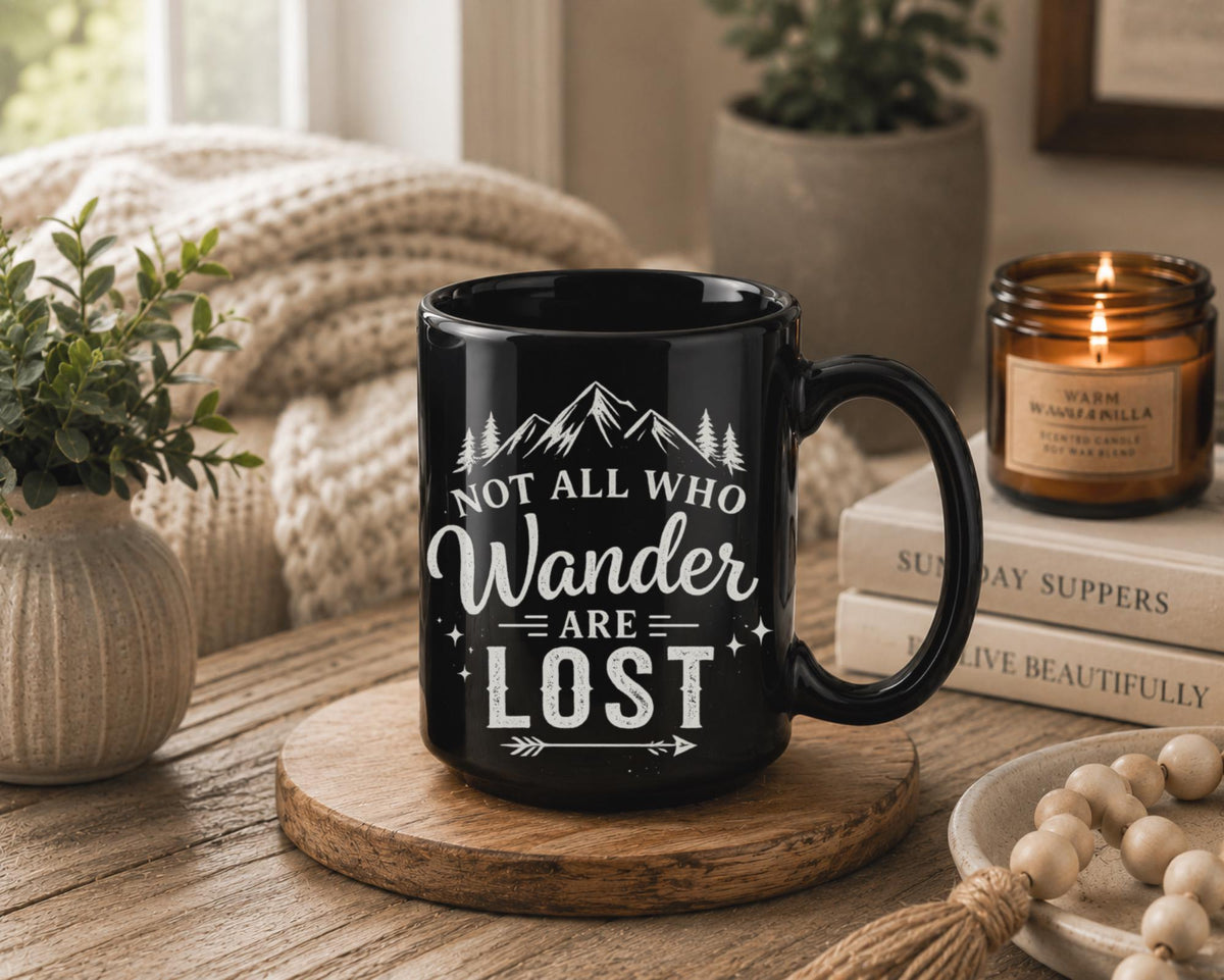 Black ceramic mug featuring mountain artwork and the phrase "Not All Who Wander Are Lost" displayed in white typography on a cozy rustic background.