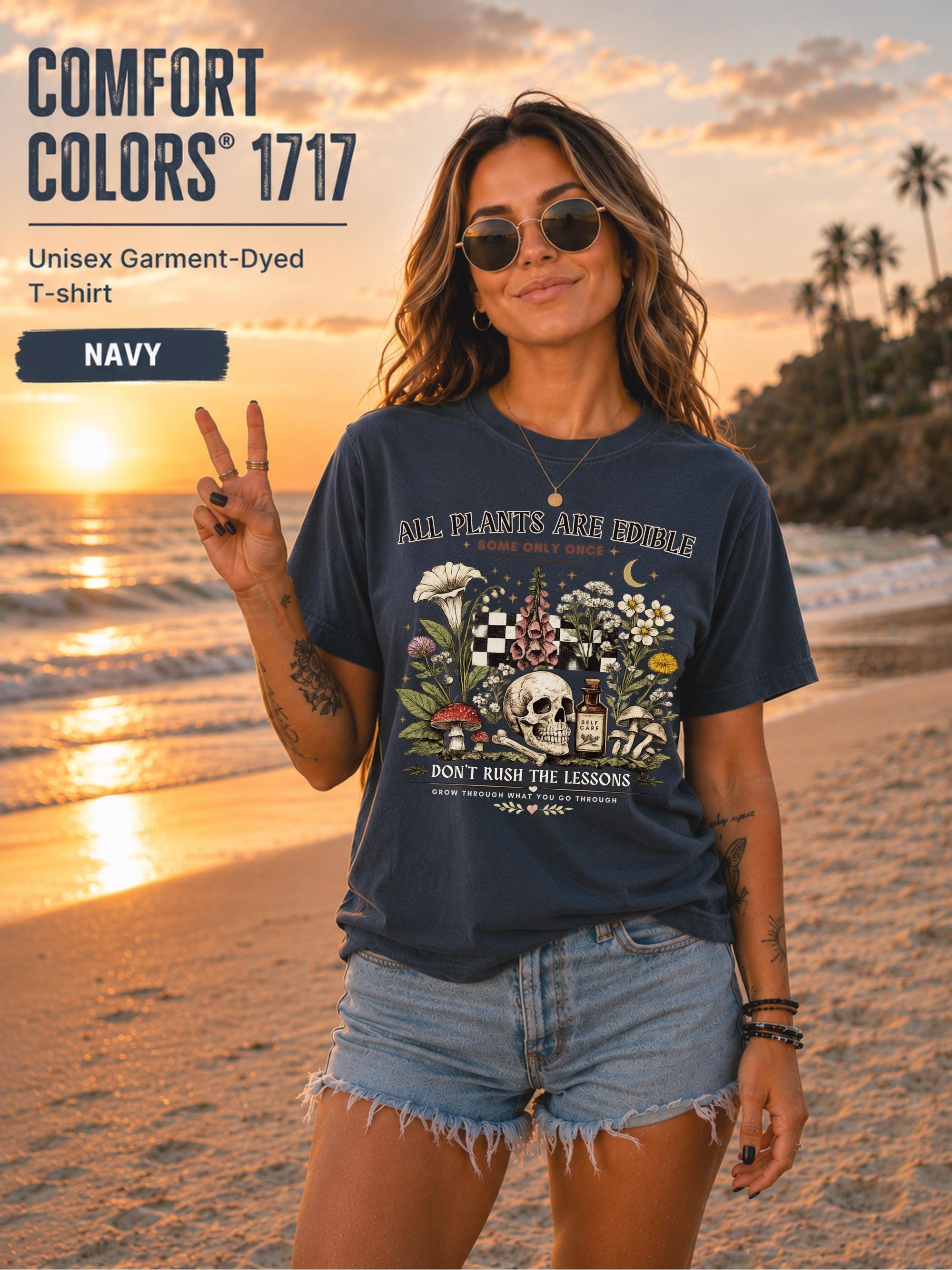 Woman wearing a navy Comfort Colors 1717 garment-dyed T-shirt featuring a vintage botanical skull design with mushrooms, flowers, and the quote "All Plants Are Edible... Some Only Once" on a beach at sunset.
