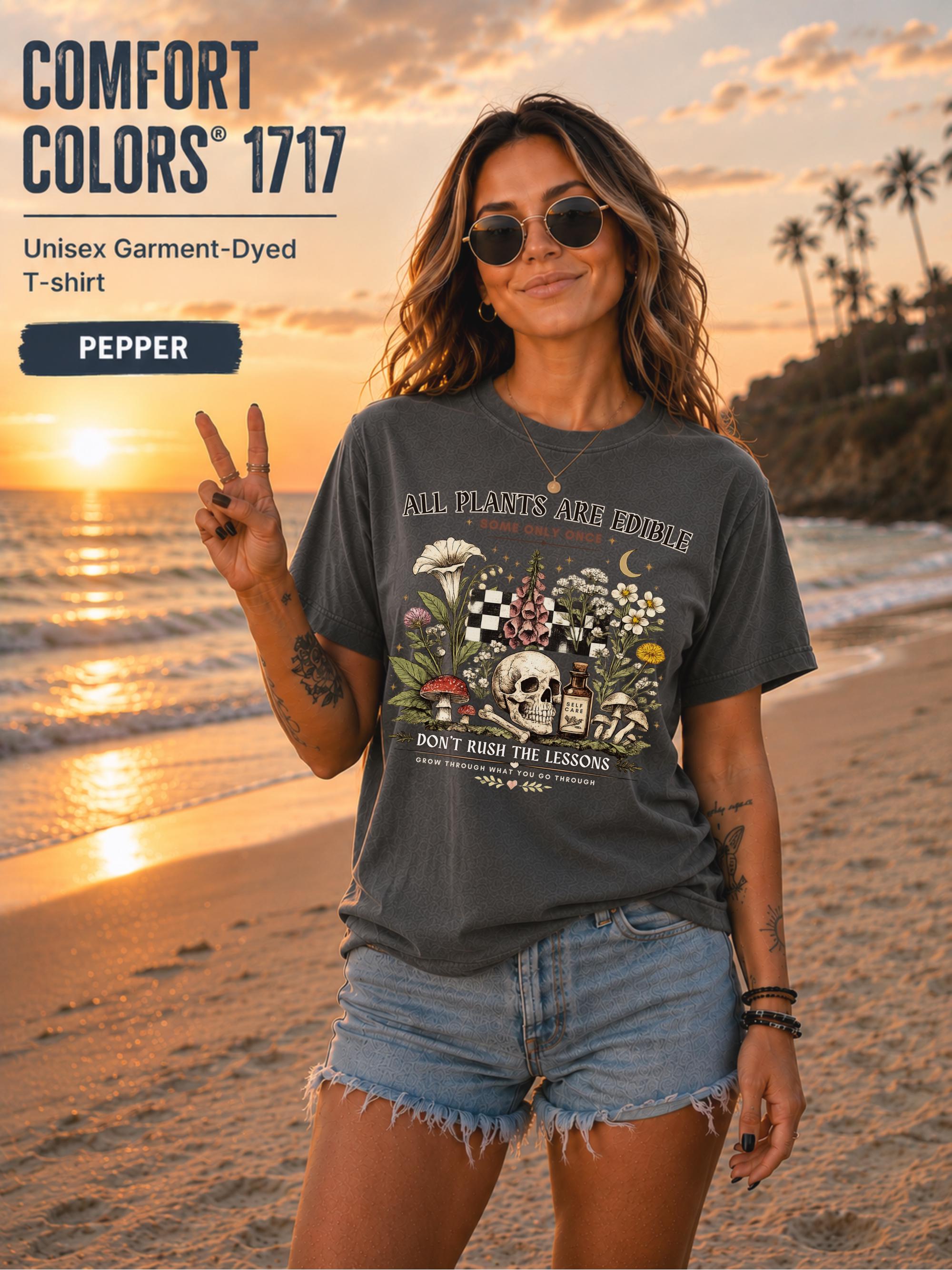 Woman wearing a pepper Comfort Colors 1717 garment-dyed T-shirt featuring a vintage botanical skull design with mushrooms, flowers, and the quote "All Plants Are Edible... Some Only Once" on a beach at sunset.
