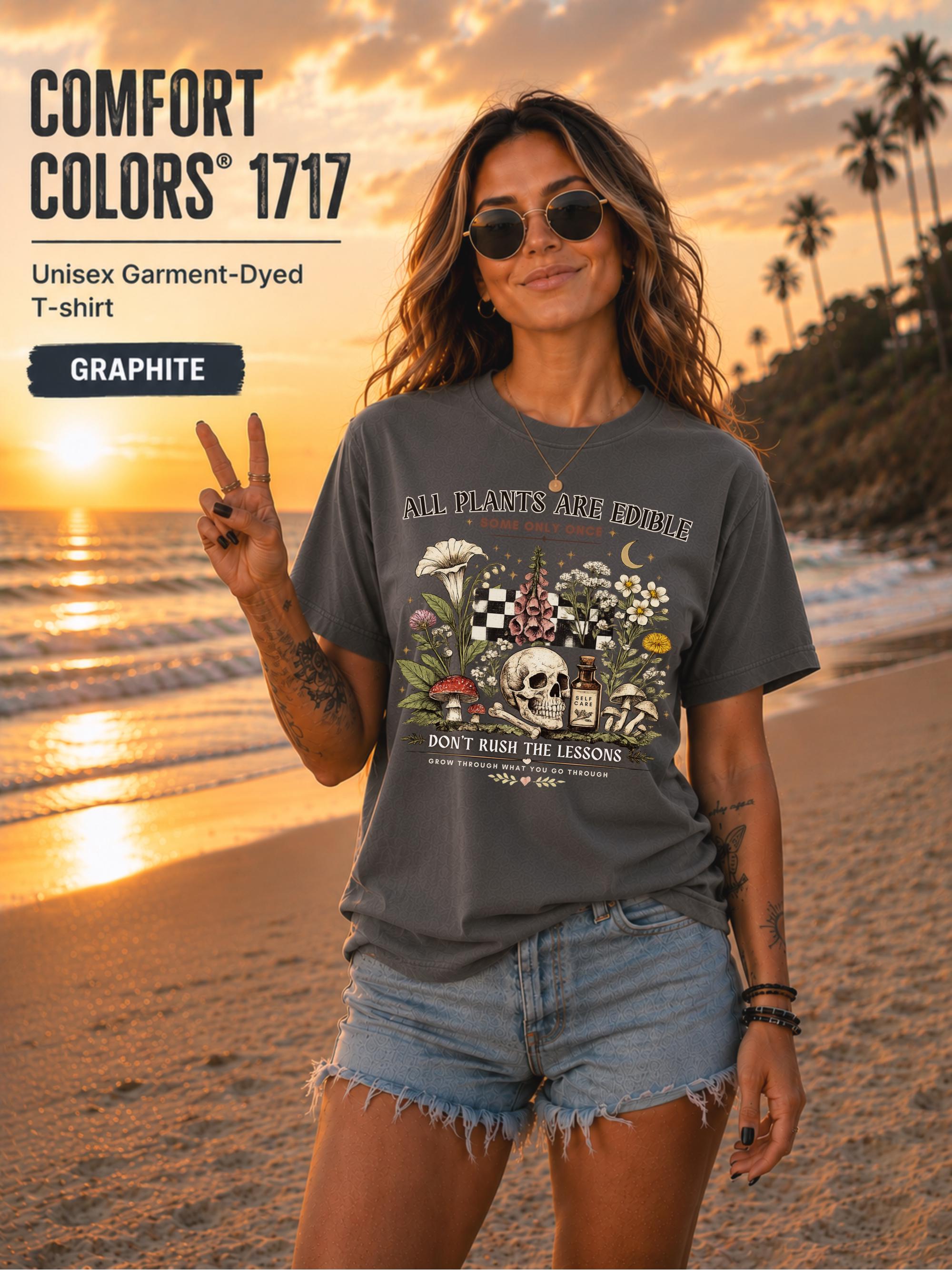 Woman wearing a graphite Comfort Colors 1717 garment-dyed T-shirt featuring a vintage botanical skull design with mushrooms, flowers, and the quote "All Plants Are Edible... Some Only Once" on a beach at sunset.
