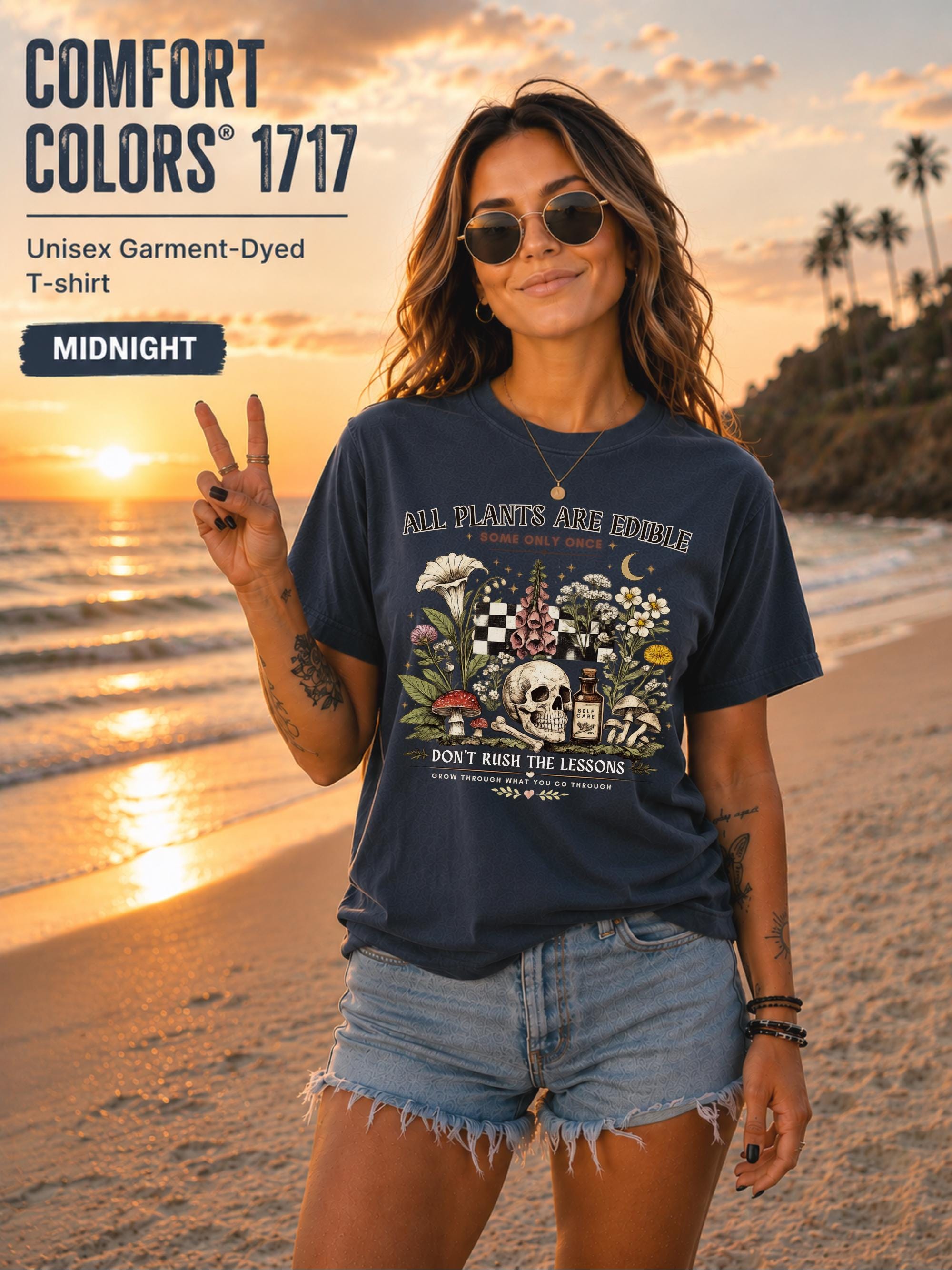 Woman wearing a midnight Comfort Colors 1717 garment-dyed T-shirt featuring a vintage botanical skull design with mushrooms, flowers, and the quote "All Plants Are Edible... Some Only Once" on a beach at sunset.