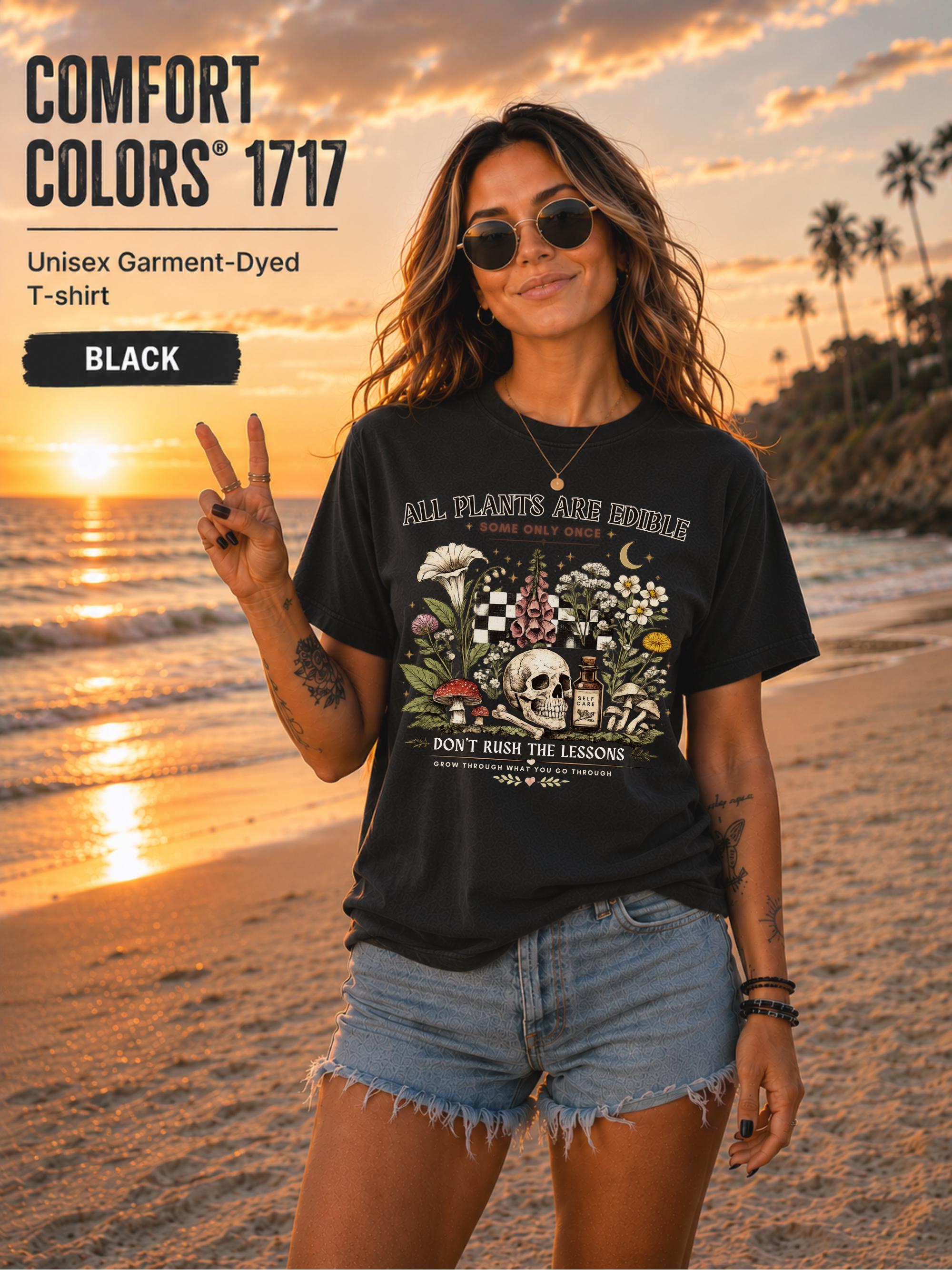 Woman wearing a black Comfort Colors 1717 garment-dyed T-shirt featuring a vintage botanical skull design with mushrooms, flowers, and the quote "All Plants Are Edible... Some Only Once" on a beach at sunset.