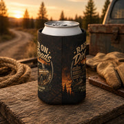 Regular can cooler with "Raised on Dirt Roads" vintage truck design displayed on a rustic wooden tailgate beside work gloves and rope at sunset, ideal for country, camping, and outdoor enthusiasts.