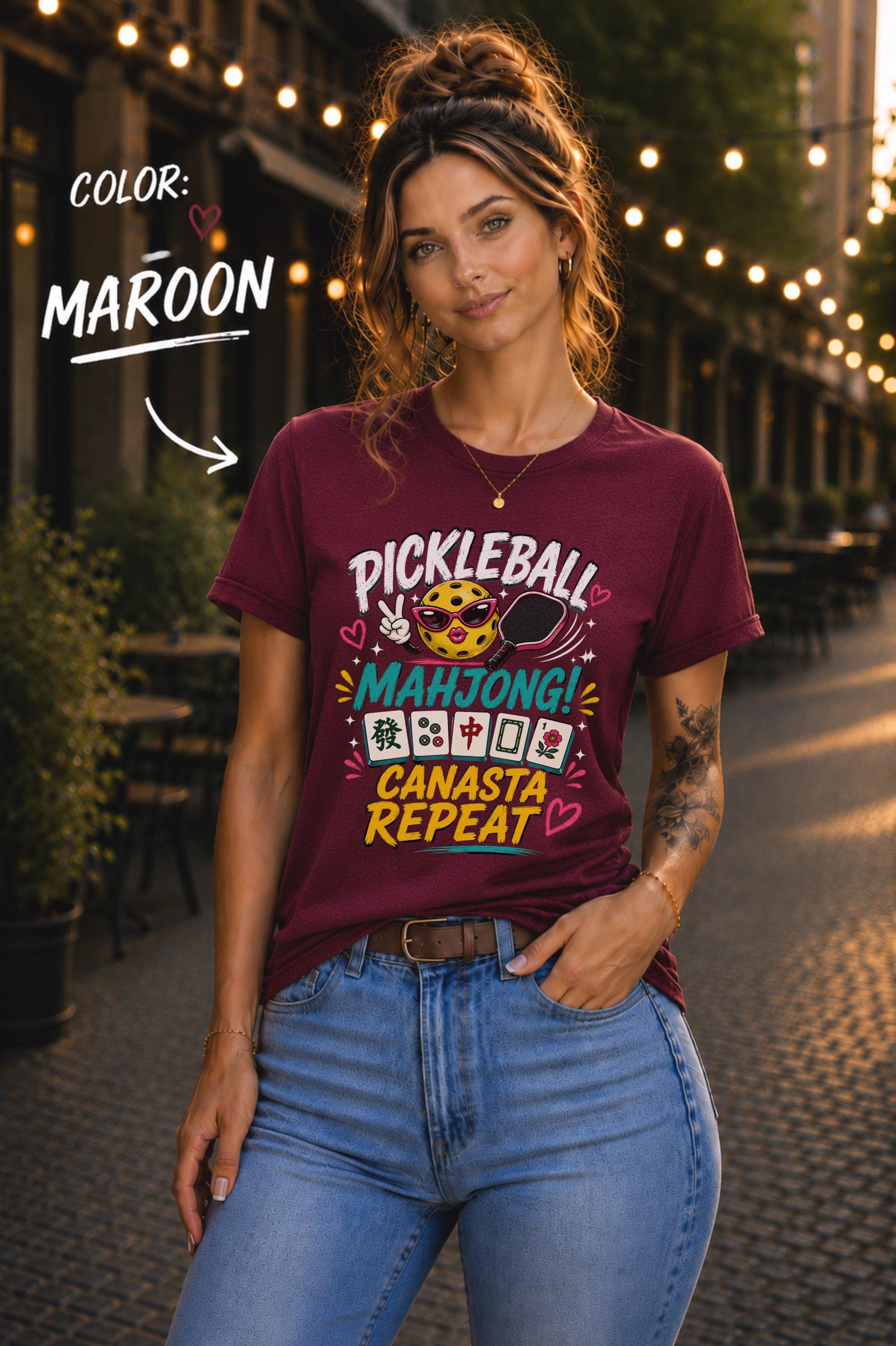 Woman wearing a maroon graphic t-shirt featuring the colorful phrase "Pickleball Mahjong Canasta Repeat" with a smiling pickleball character, paddle, and Mahjong tiles, styled for casual everyday wear.
