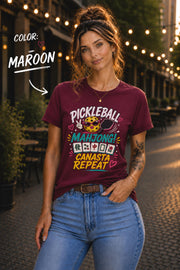 Woman wearing a maroon graphic t-shirt featuring the colorful phrase "Pickleball Mahjong Canasta Repeat" with a smiling pickleball character, paddle, and Mahjong tiles, styled for casual everyday wear.