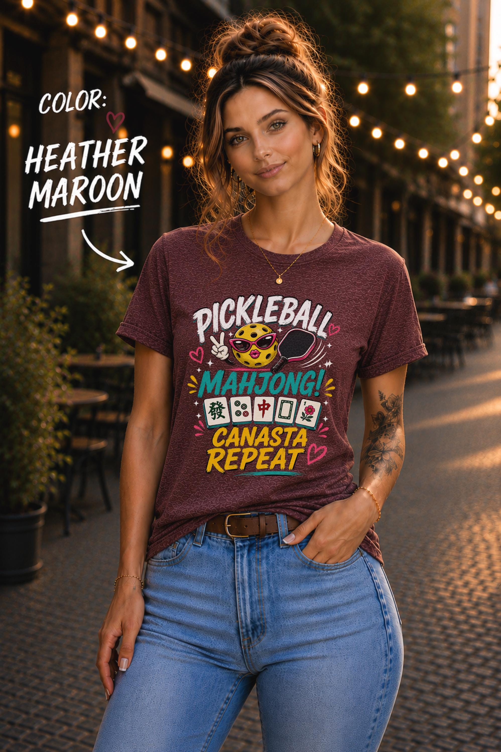 Woman wearing a heather maroon graphic t-shirt featuring the colorful phrase "Pickleball Mahjong Canasta Repeat" with a smiling pickleball character, paddle, and Mahjong tiles, styled for casual everyday wear.