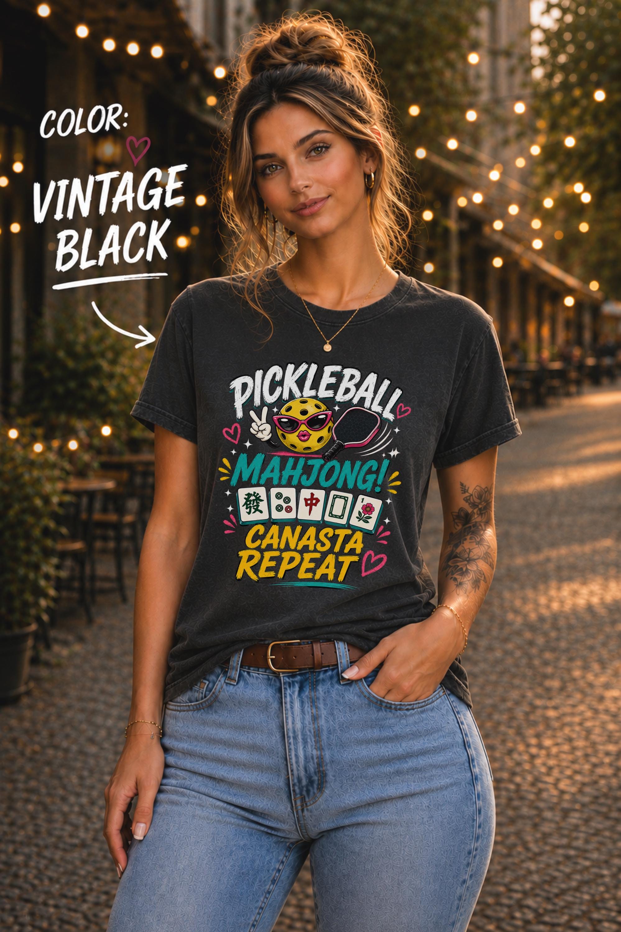 Woman wearing a vintage black graphic t-shirt featuring the colorful phrase "Pickleball Mahjong Canasta Repeat" with a smiling pickleball character, paddle, and Mahjong tiles, styled for casual everyday wear.