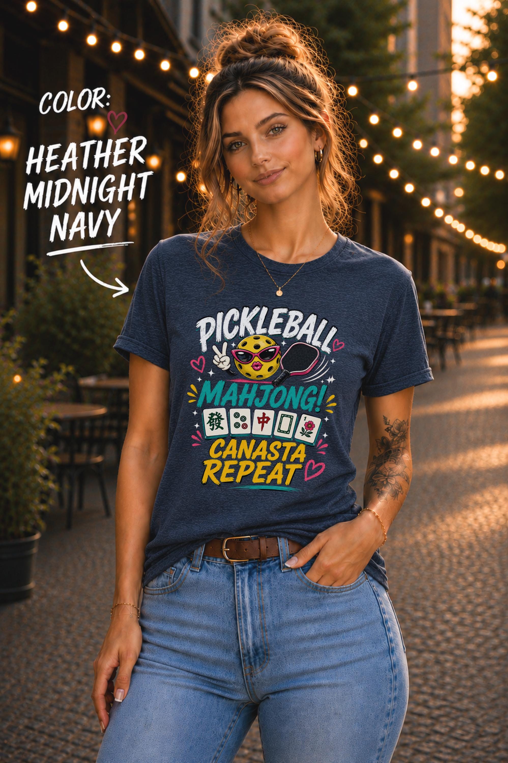 Woman wearing a heather midnight navy graphic t-shirt featuring the colorful phrase "Pickleball Mahjong Canasta Repeat" with a smiling pickleball character, paddle, and Mahjong tiles, styled for casual everyday wear.