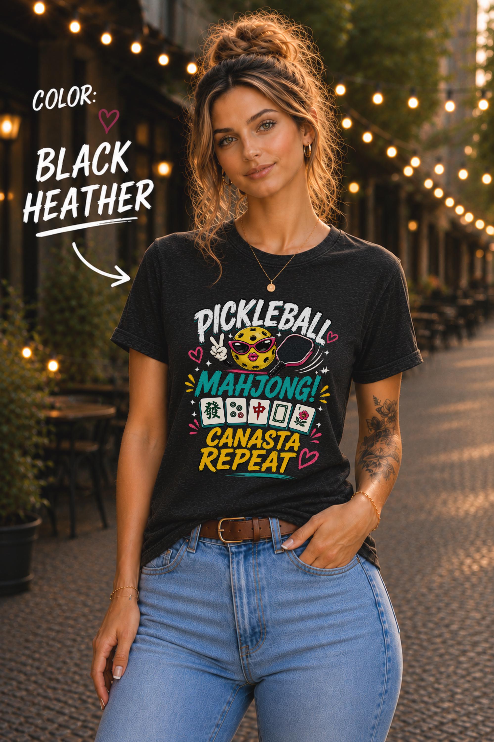 Woman wearing a black heather graphic t-shirt featuring the colorful phrase "Pickleball Mahjong Canasta Repeat" with a smiling pickleball character, paddle, and Mahjong tiles, styled for casual everyday wear.