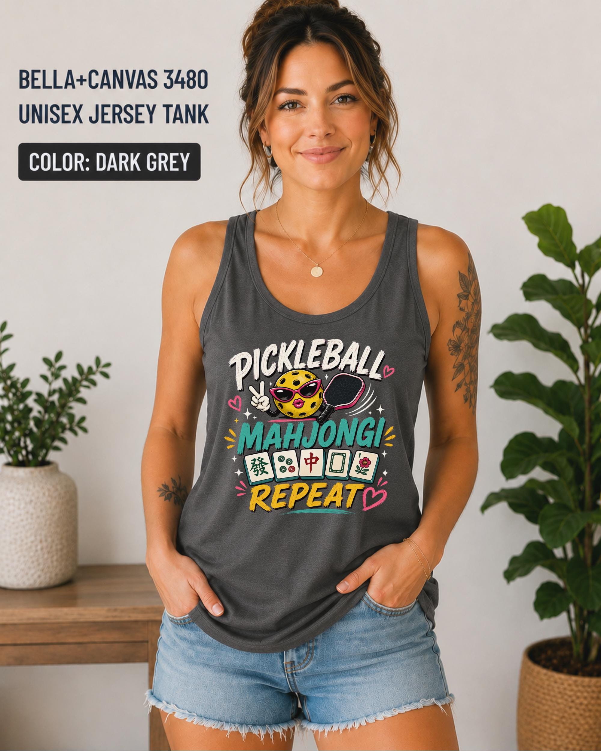 Woman wearing a dark grey Bella+Canvas 3480 unisex jersey tank featuring a colorful "Pickleball Mahjong Repeat" graphic with a smiling pickleball, paddle, Mahjong tiles, and bright playful lettering.