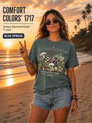 Woman wearing a blue spruce Comfort Colors 1717 garment-dyed T-shirt featuring a vintage botanical skull design with mushrooms, flowers, and the quote "All Plants Are Edible... Some Only Once" on a beach at sunset.