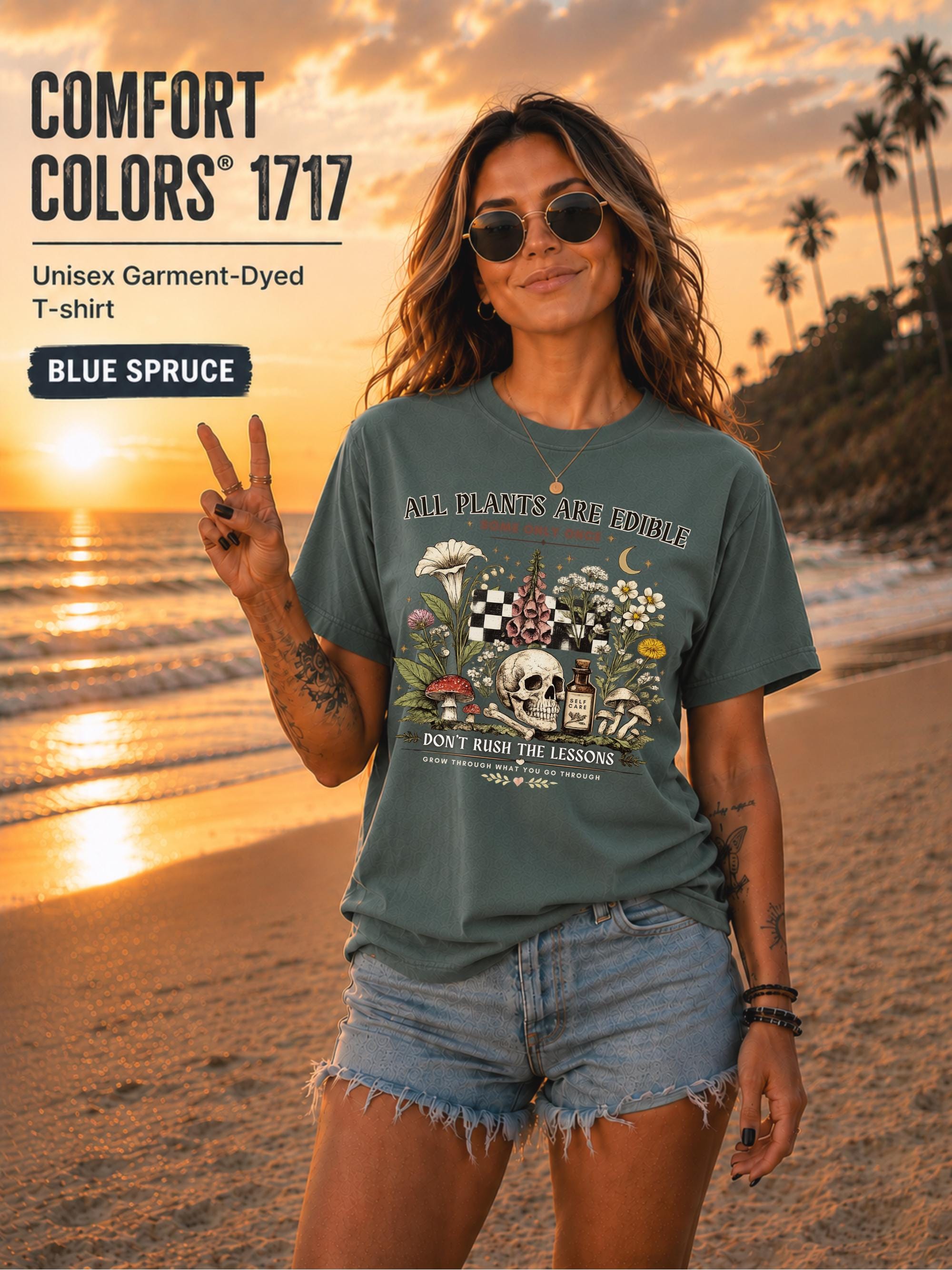 Woman wearing a blue spruce Comfort Colors 1717 garment-dyed T-shirt featuring a vintage botanical skull design with mushrooms, flowers, and the quote "All Plants Are Edible... Some Only Once" on a beach at sunset.