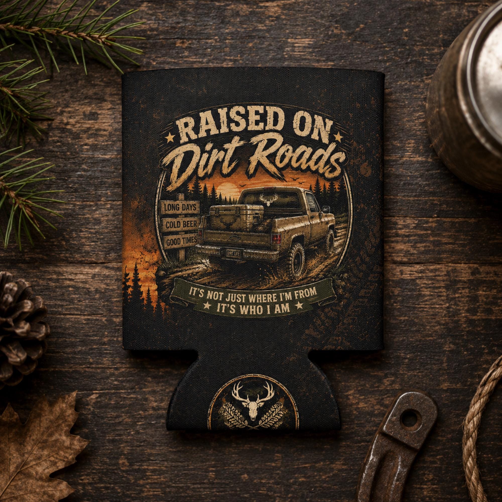 Regular can cooler with "Raised on Dirt Roads" vintage truck design displayed on a rustic wooden tailgate beside work gloves and rope at sunset, ideal for country, camping, and outdoor enthusiasts.