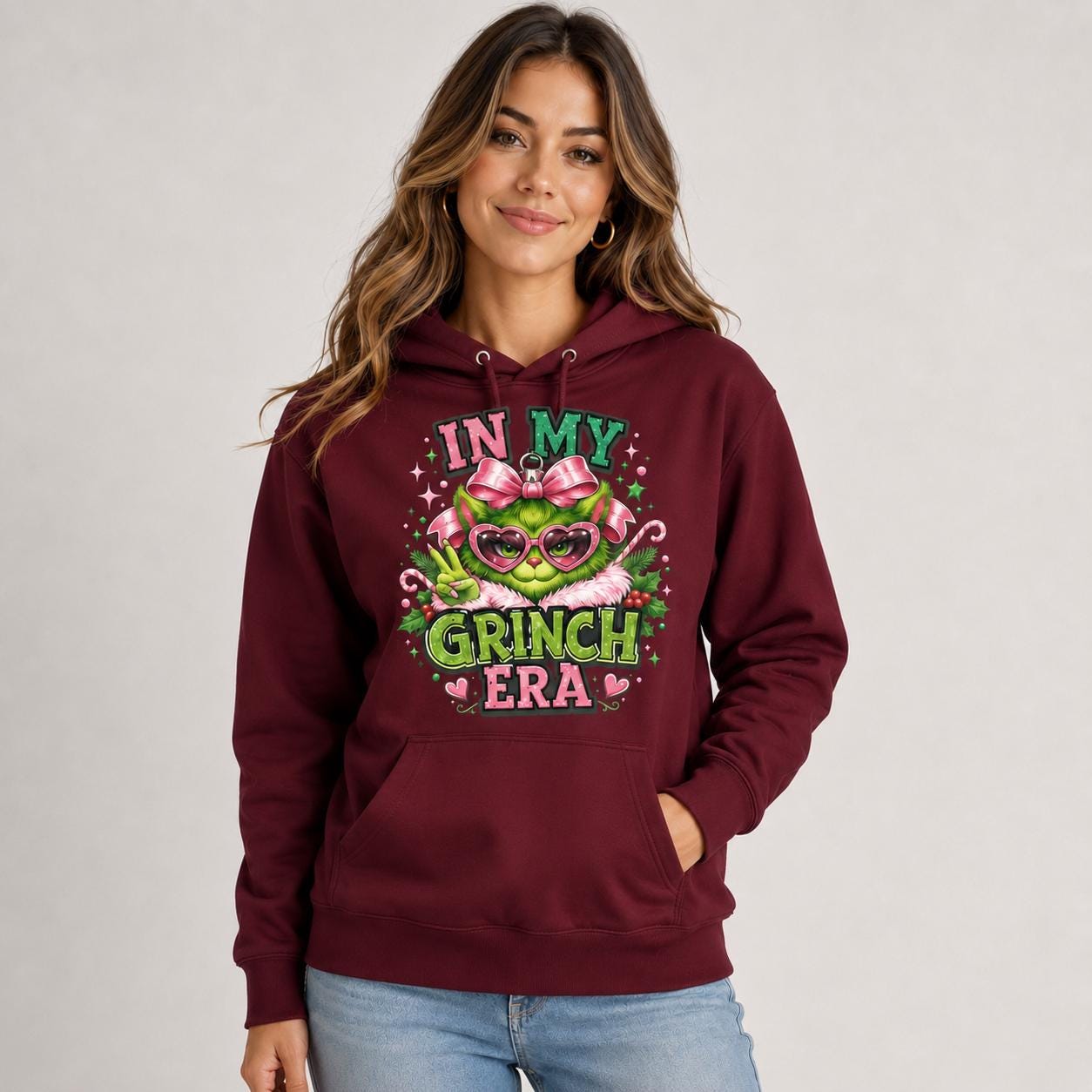Female model wearing a maroon holiday hoodie with a pink and green festive character design printed on the front.