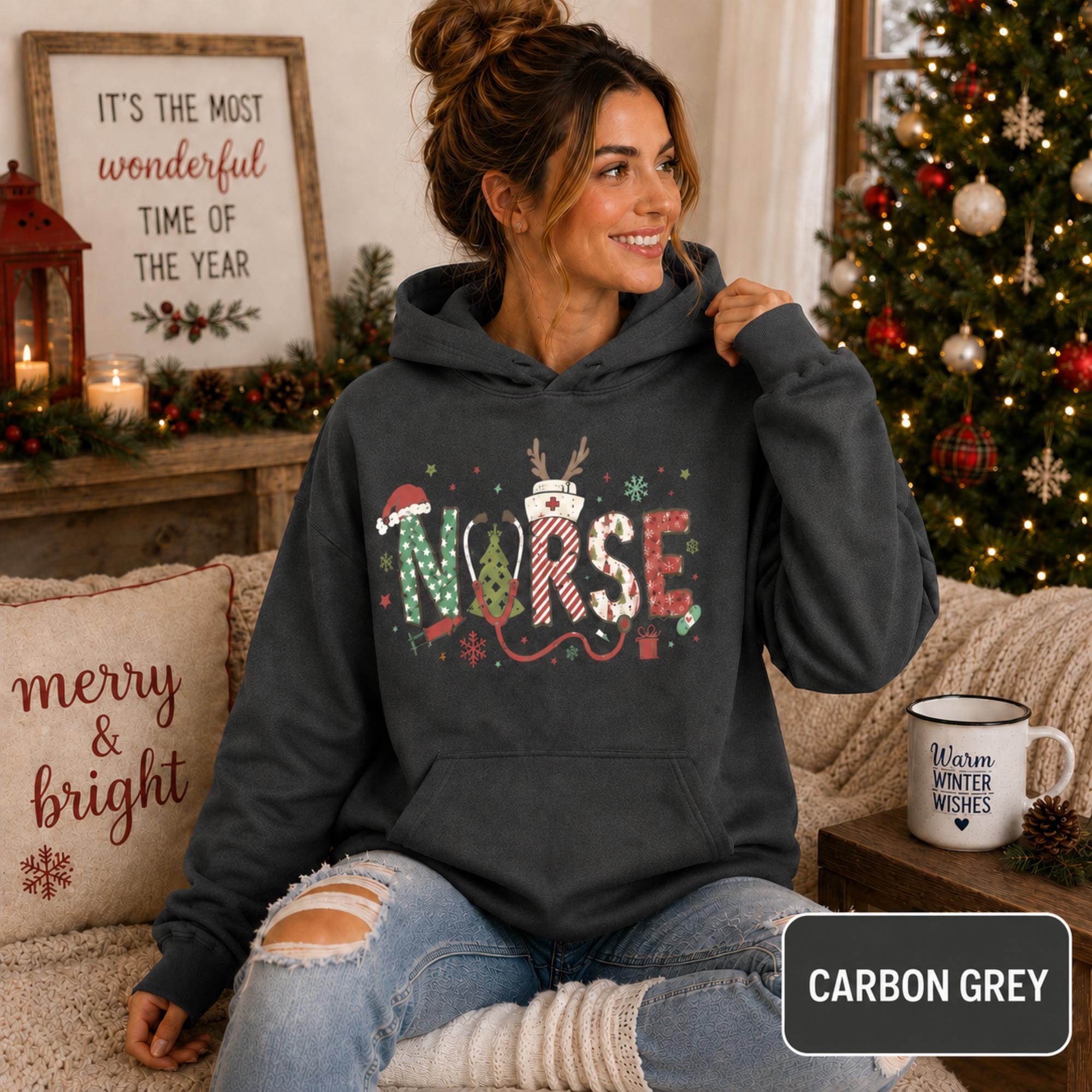 Female model wearing a carbon grey Christmas nurse hoodie with colorful festive NURSE lettering in a cozy holiday living room.