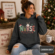 Female model wearing a carbon grey Christmas nurse hoodie with colorful festive NURSE lettering in a cozy holiday living room.