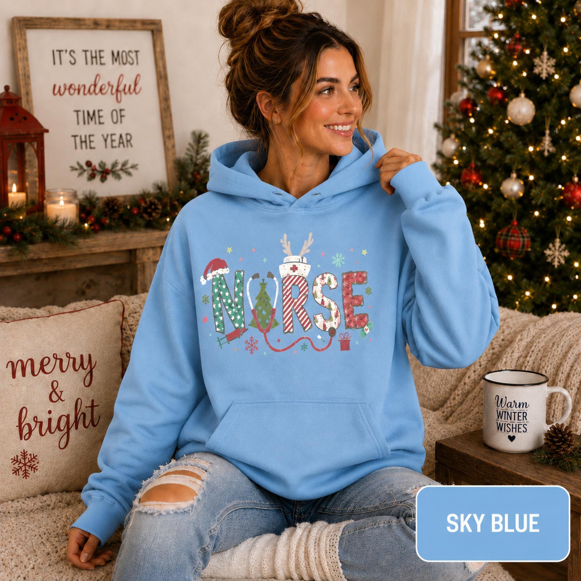 Female model wearing a sky blue Christmas nurse hoodie with colorful festive NURSE lettering in a cozy holiday living room.