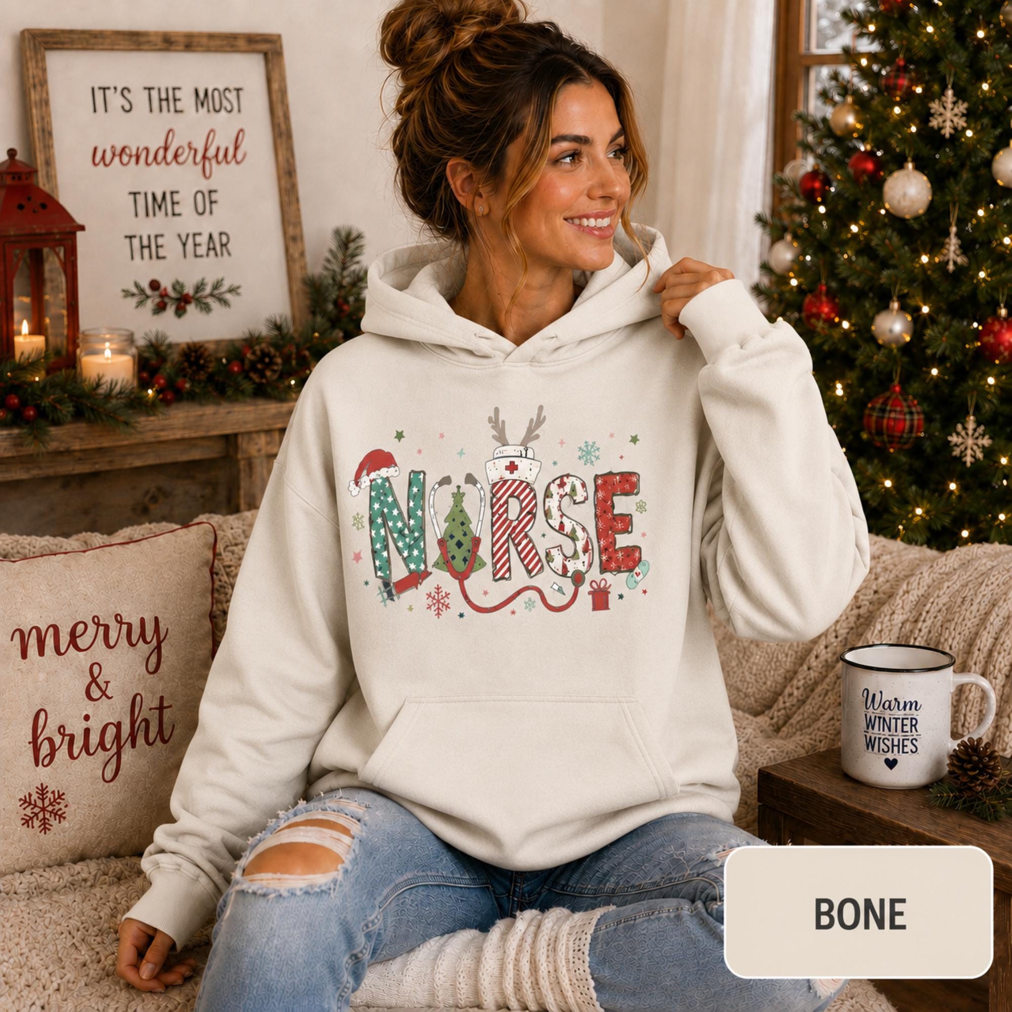 Female model wearing a bone Christmas nurse hoodie with colorful festive NURSE lettering in a cozy holiday living room.