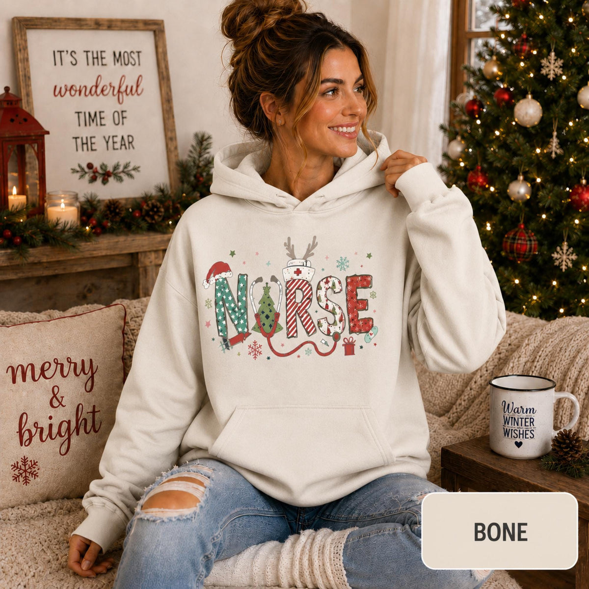 Female model wearing a bone Christmas nurse hoodie with colorful festive NURSE lettering in a cozy holiday living room.