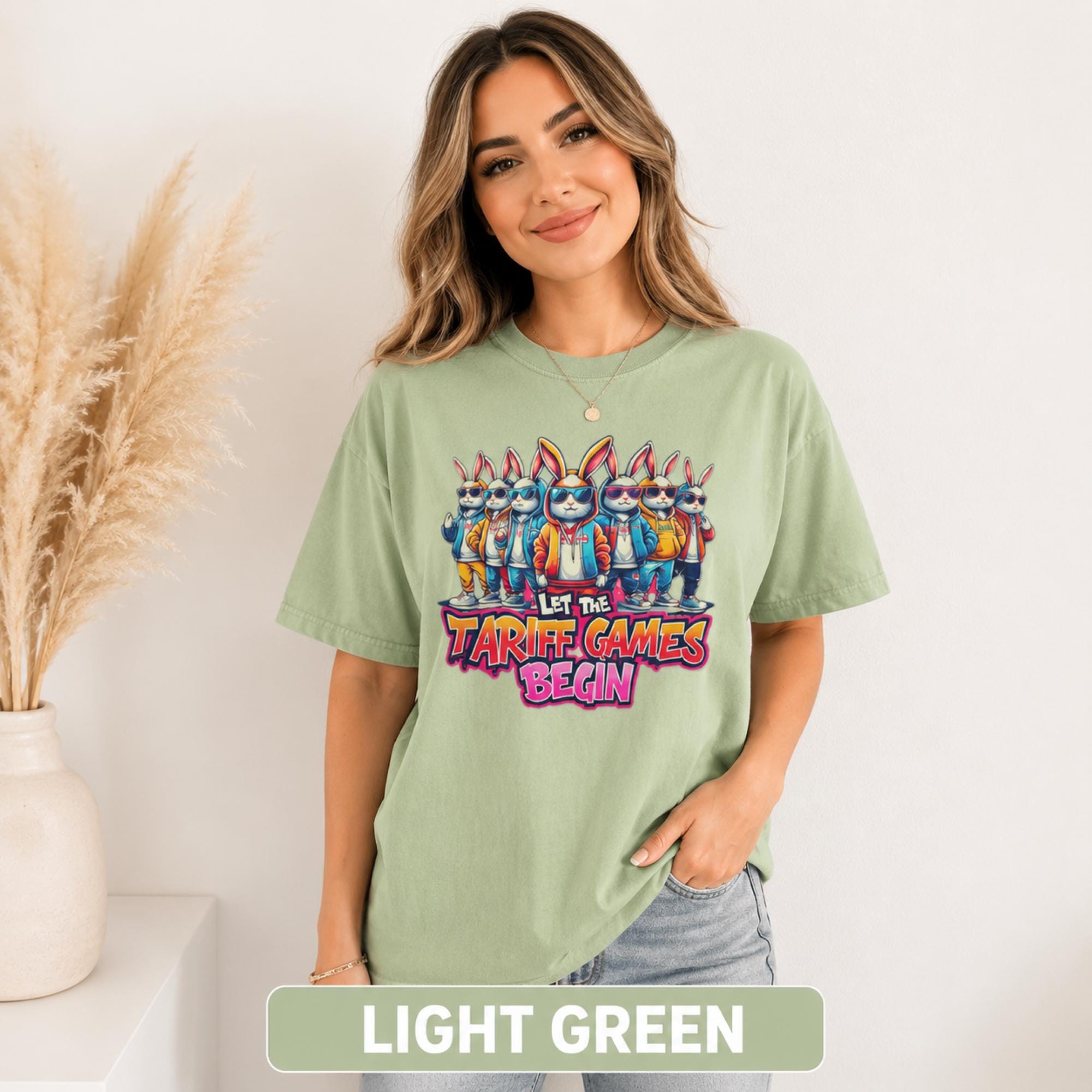 Female model wearing a light green T-shirt with colorful cartoon rabbits and “Let the Tariff Games Begin” graphic design.