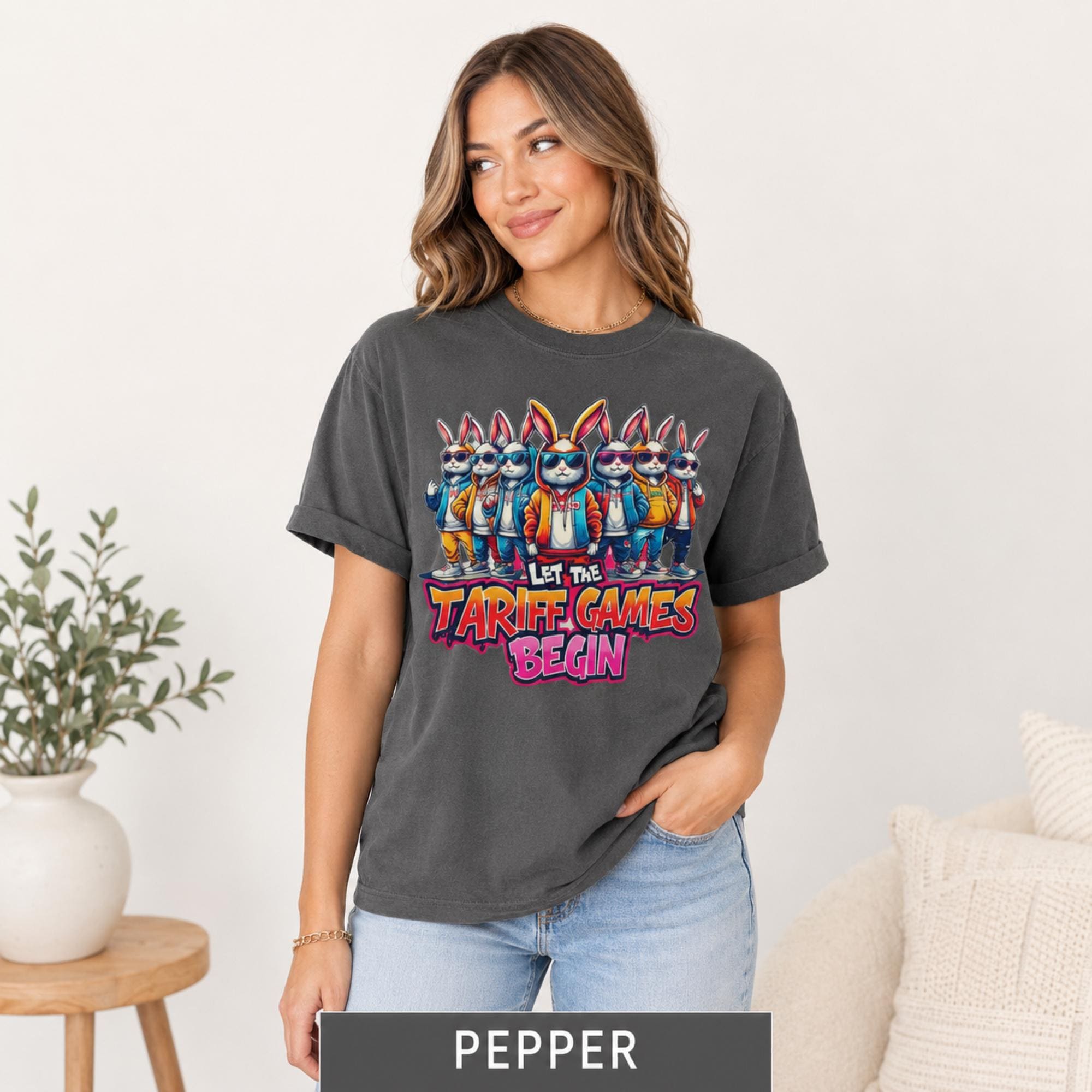Female model wearing a pepper grey T-shirt featuring cartoon rabbits in sunglasses and “Let the Tariff Games Begin” design.